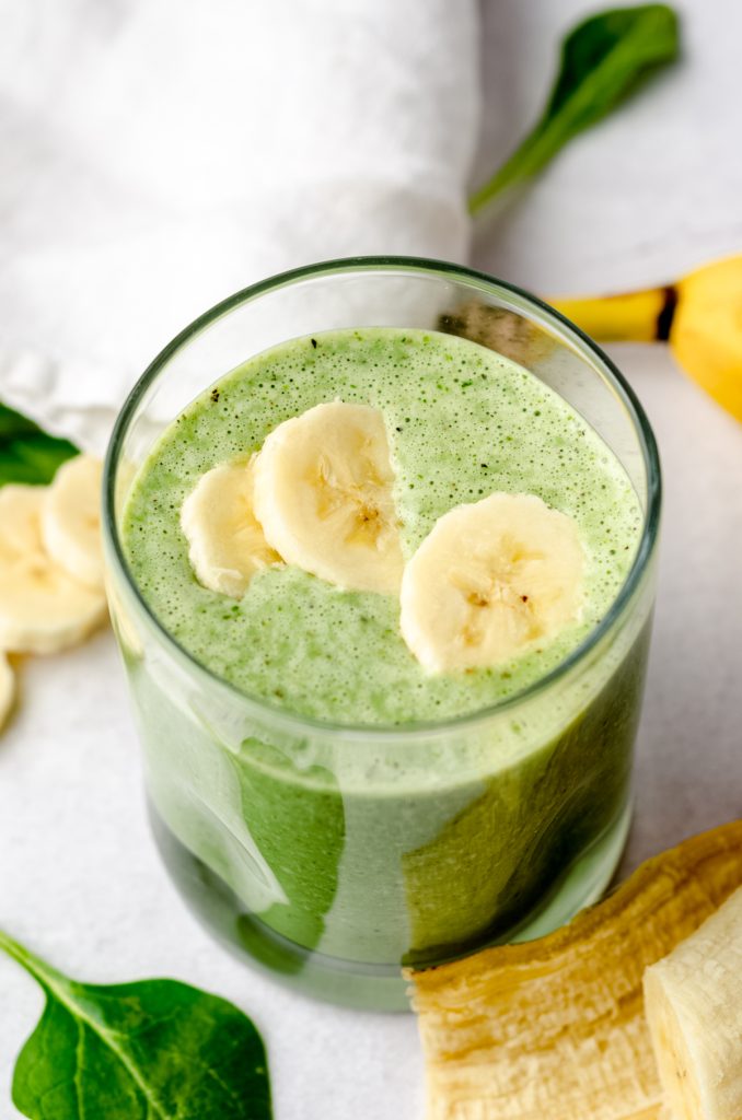 A glass with a green breakfast smoothie in it on a surface with bananas and spinach in the foreground and background and banana slices on top of the smoothie.