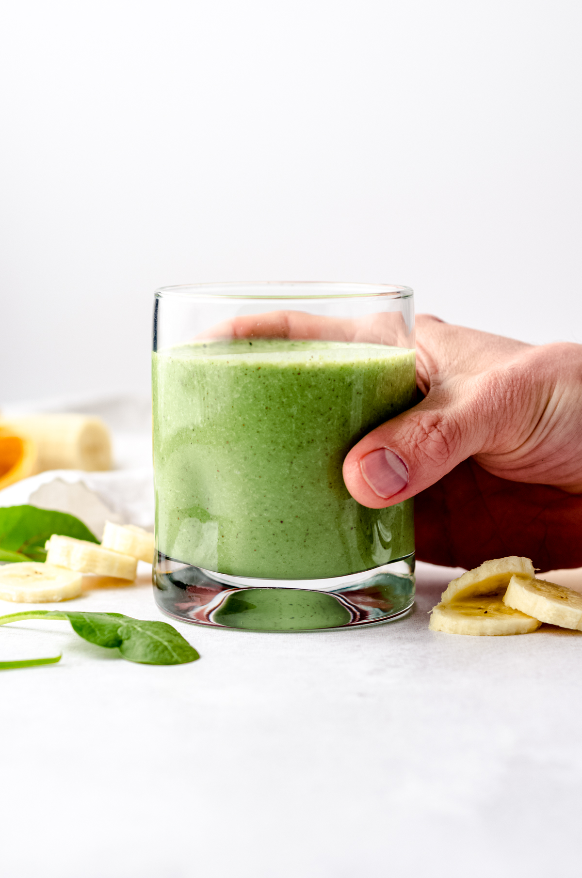 A glass with a green breakfast smoothie in it on a surface with bananas and spinach in the foreground and background and someone has their hand around the glass.
