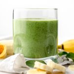 A glass with a green breakfast smoothie in it on a surface with bananas and spinach in the foreground and background.