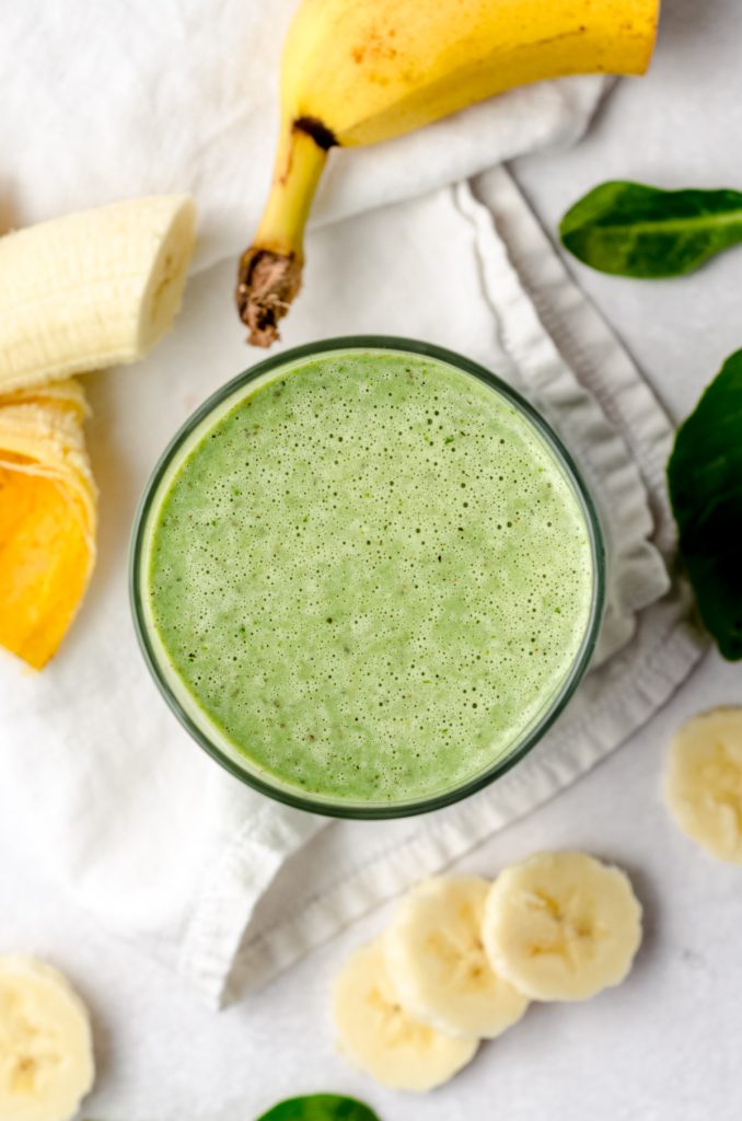 Aerial photo of a green breakfast smoothie on a surface with bananas and spinach around it.