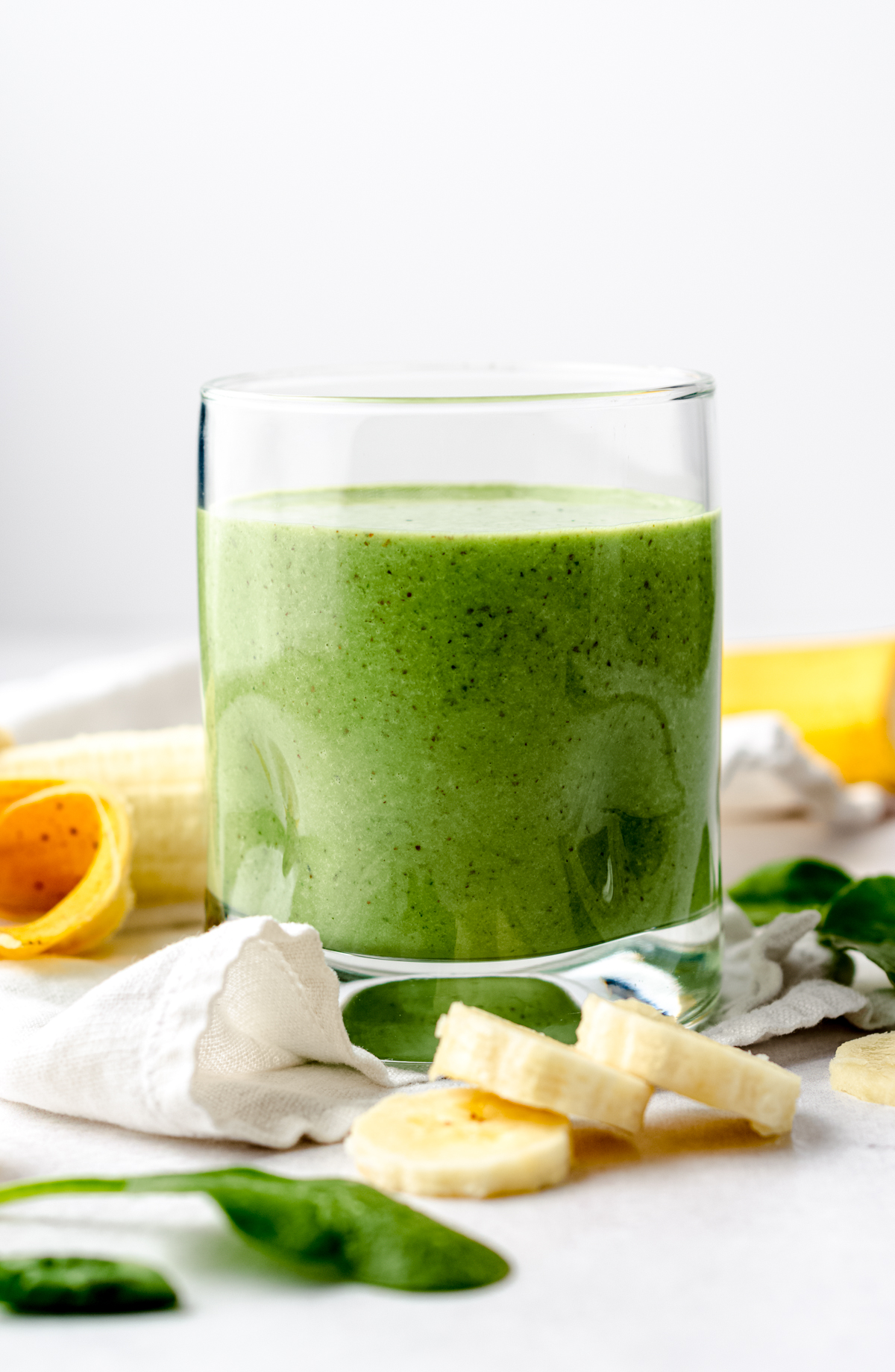 A glass with a green breakfast smoothie in it on a surface with bananas and spinach in the foreground and background.