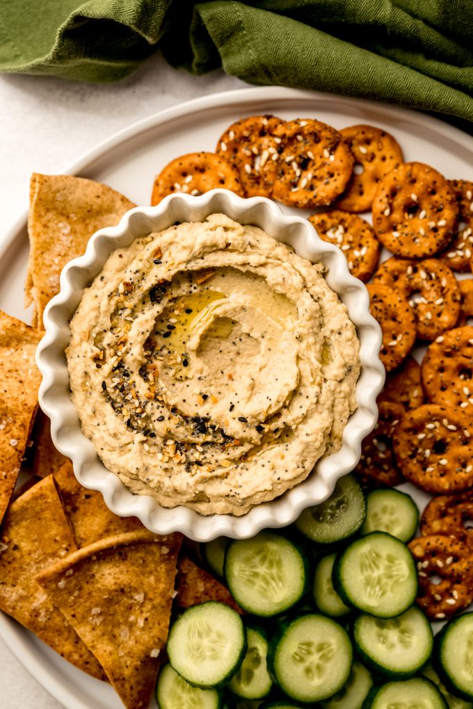 Aerial photo of a plate of crackers, pretzels, and cucumber slices with a bowl of everything bagel hummus on it.