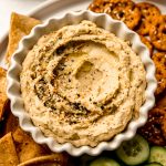 Aerial photo of a plate of crackers, pretzels, and cucumber slices with a bowl of everything bagel hummus on it.