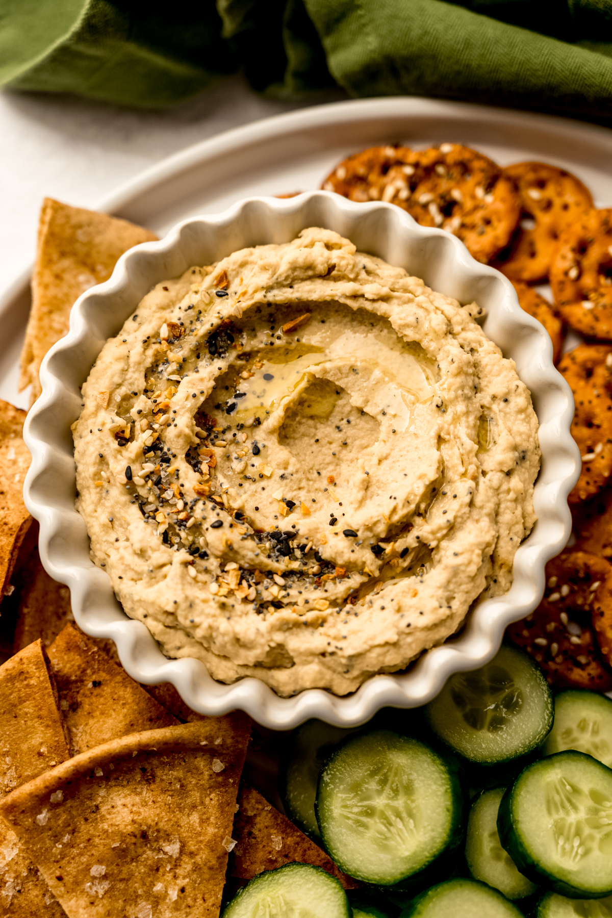 Aerial photo of a plate of crackers, pretzels, and cucumber slices with a bowl of everything bagel hummus on it.