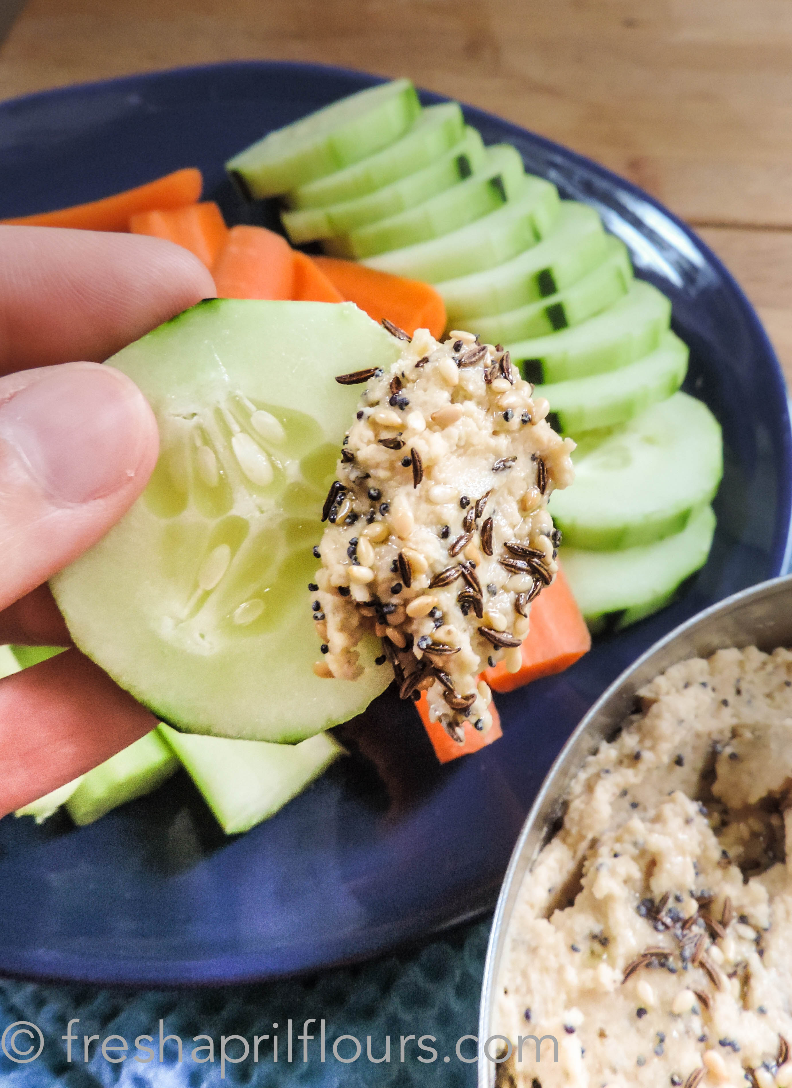 fingers holding a slice of cucumber dipped into everything bagel hummus