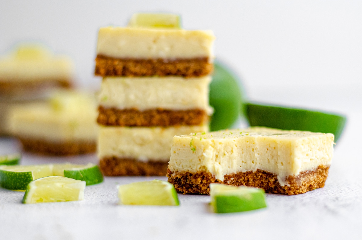 a key lime pie bar in the foreground with a bite taken out of it and a stack of key lime pie bars in the background