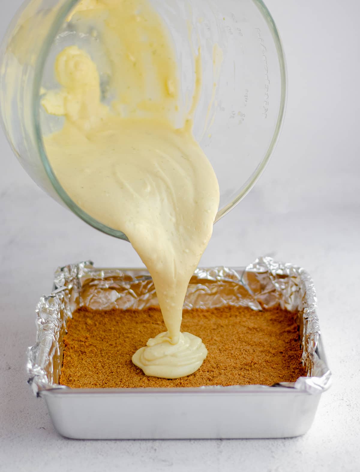 key lime pie batter being poured onto a baked graham cracker crust in a foil lined baking pan