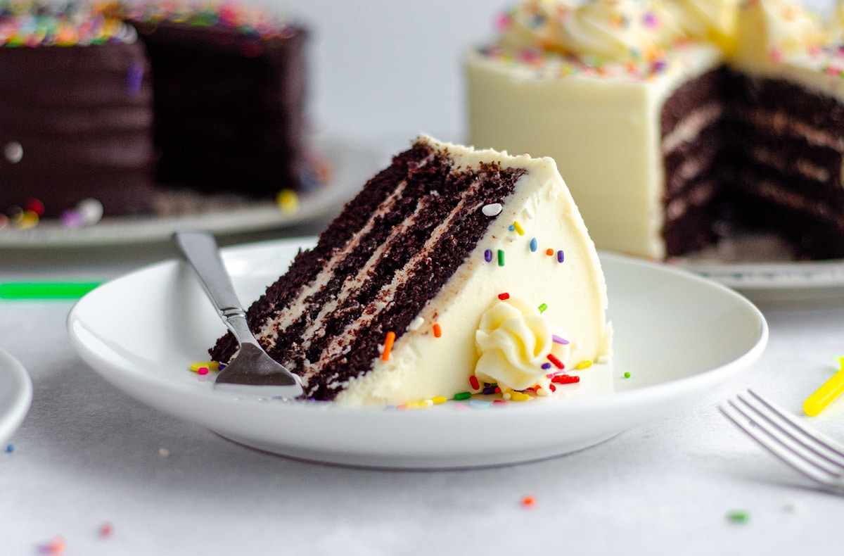 slice of chocolate layer cake with vanilla buttercream and rainbow sprinkles on a white plate with a fork