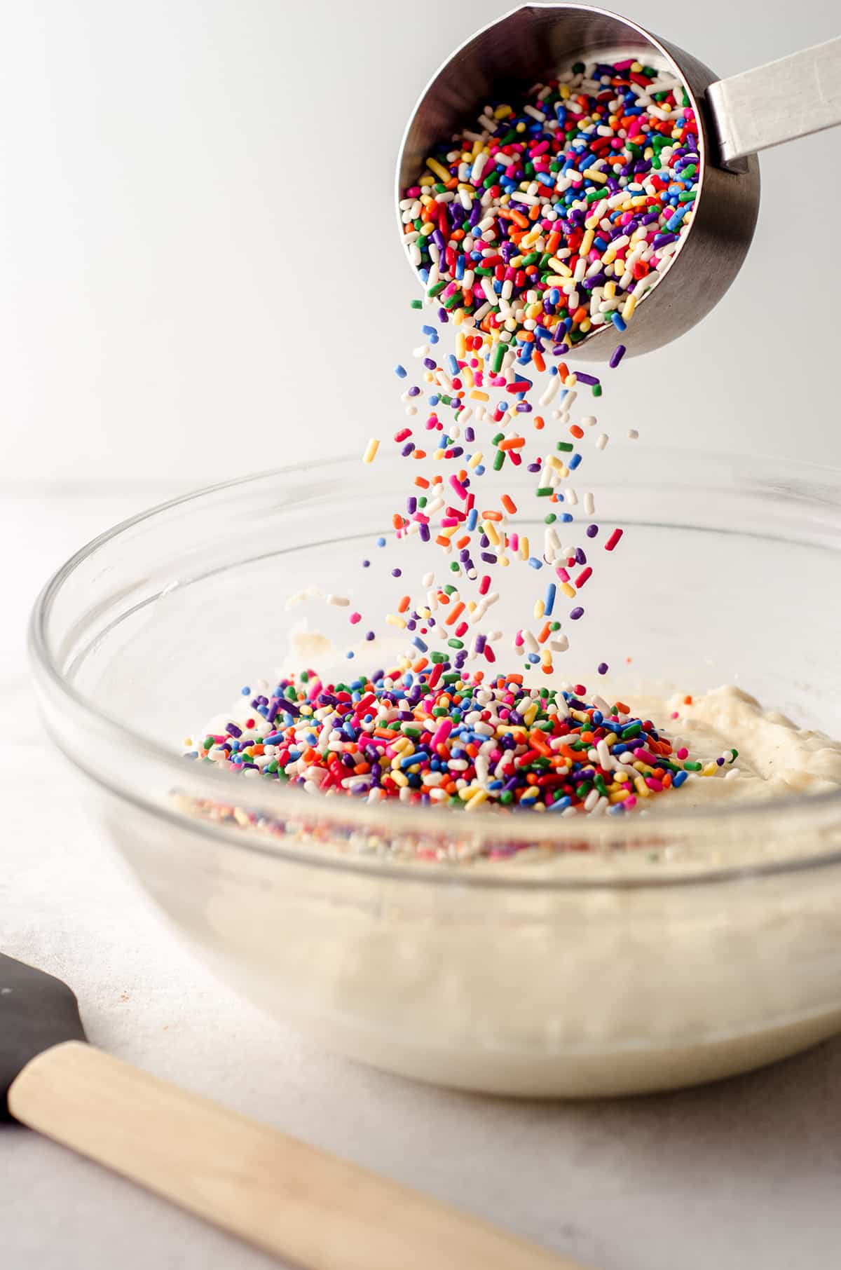 pouring rainbow sprinkles into funfetti donut batter in a glass bowl