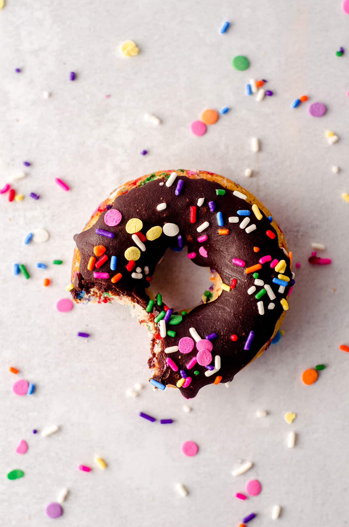 aerial photo of one funfetti donut with chocolate glaze with a bite taken out of it and surrounded by lots of rainbow sprinkles