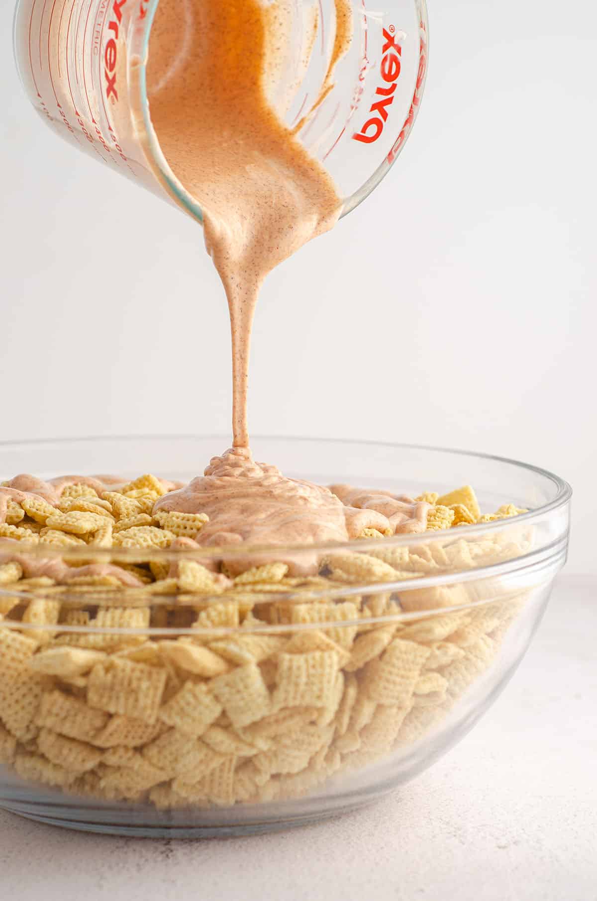 chex sitting in a glass bowl and orange candy melts being poured over them for pumpkin spice puppy chow