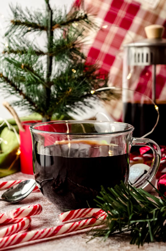 A cup of coffee sitting in a Christmas scene with a French press in the background.