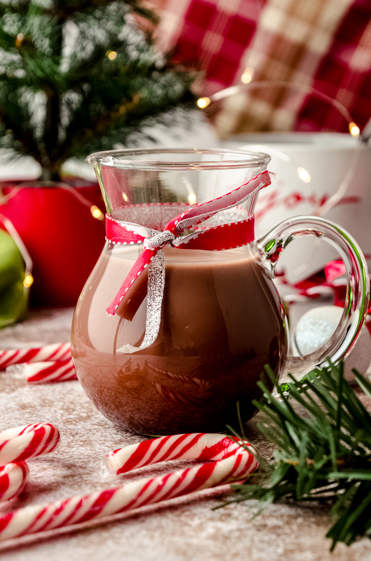 A glass carafe of peppermint mocha coffee creamer sitting on a surface surrounded by a Christmas scape.