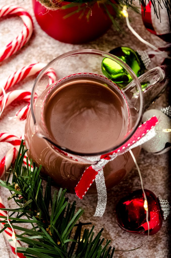 A glass carafe of peppermint mocha coffee creamer sitting on a surface surrounded by a Christmas scape.