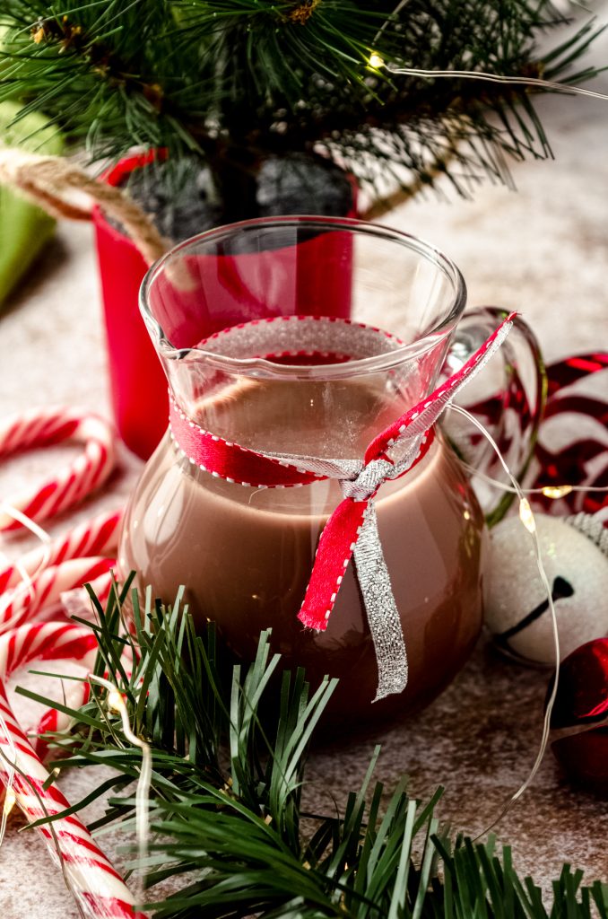 A glass carafe of peppermint mocha coffee creamer sitting on a surface surrounded by a Christmas scape.