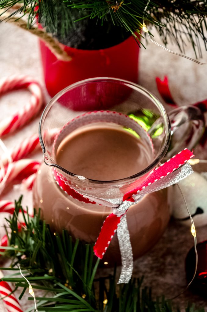 A glass carafe of peppermint mocha coffee creamer sitting on a surface surrounded by a Christmas scape.