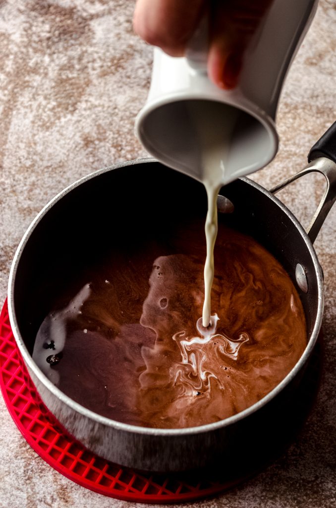 A saucepan of simple syrup with cocoa powder in it and someone is pouring a carafe of cream into it.