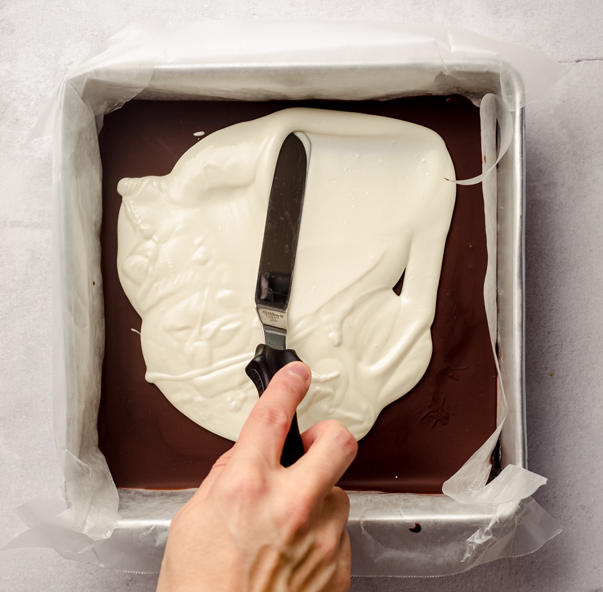 hand spreading white chocolate onto hardened semi-sweet chocolate layer to make peppermint bark