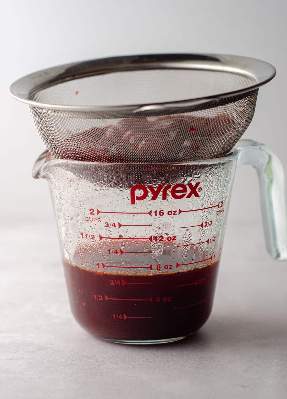 a mesh strainer sitting on top of a glass measuring cup draining macerated cranberries