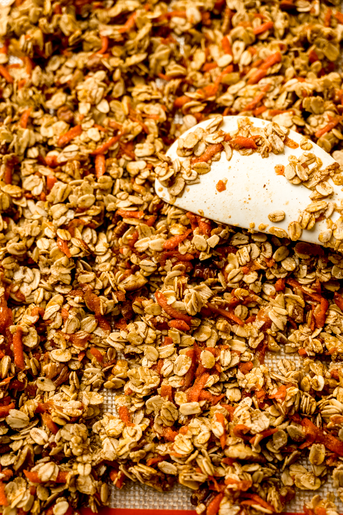 A baking sheet of carrot cake granola being spread around with a spatula.