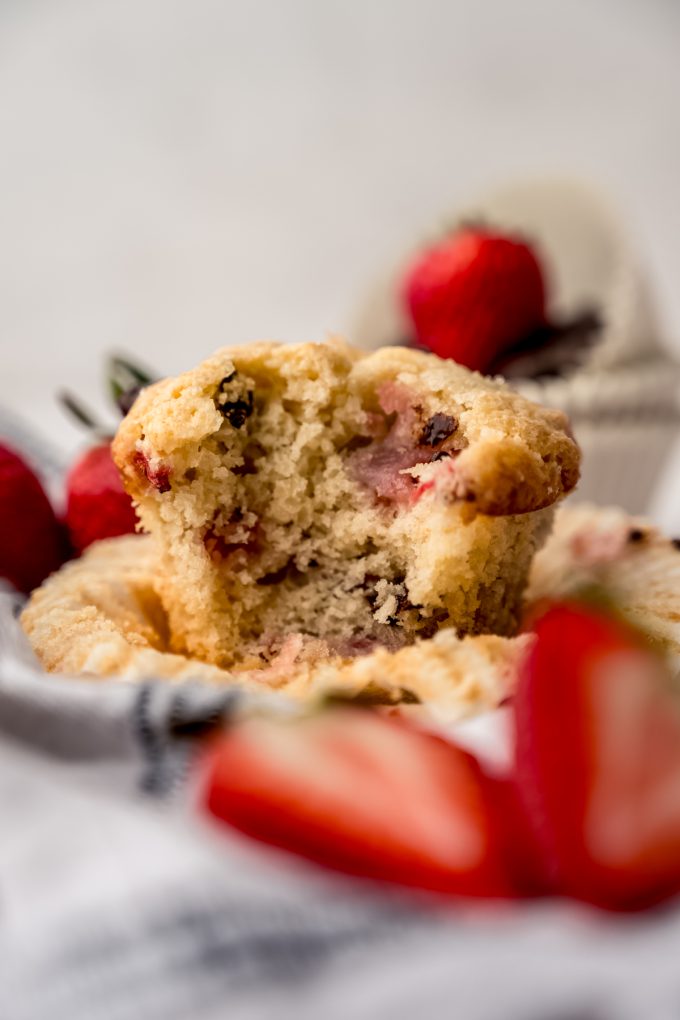 A photo of a strawberry chocolate chip muffin on a kitchen towel with strawberries around it and a bite has been taken out of it.