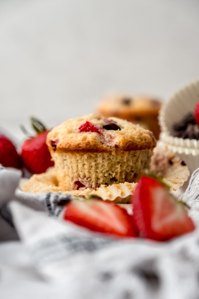 A photo of a strawberry chocolate chip muffin on a kitchen towel with strawberries around it and the wrapper has been taken off.