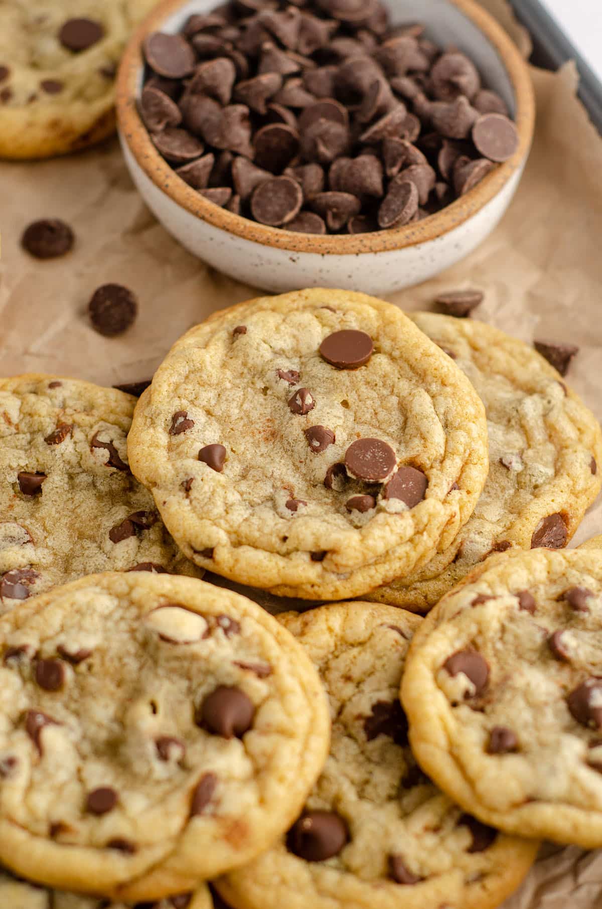 a pile of chocolate chip cookies on a baking sheet lined with parchment paper and a bowl of chocolate chips
