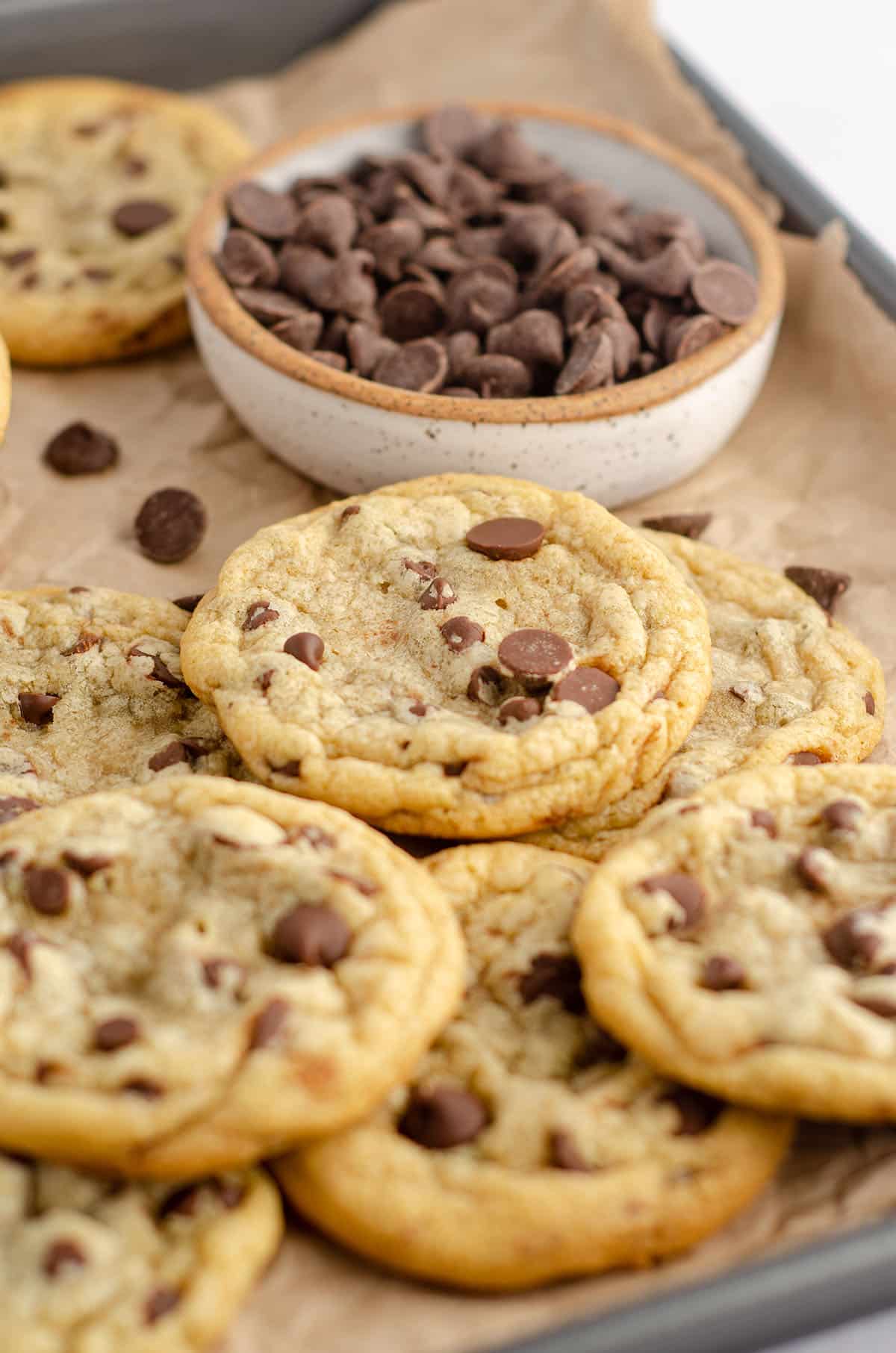 a pile of chocolate chip cookies on a baking sheet lined with parchment paper and a bowl of chocolate chips
