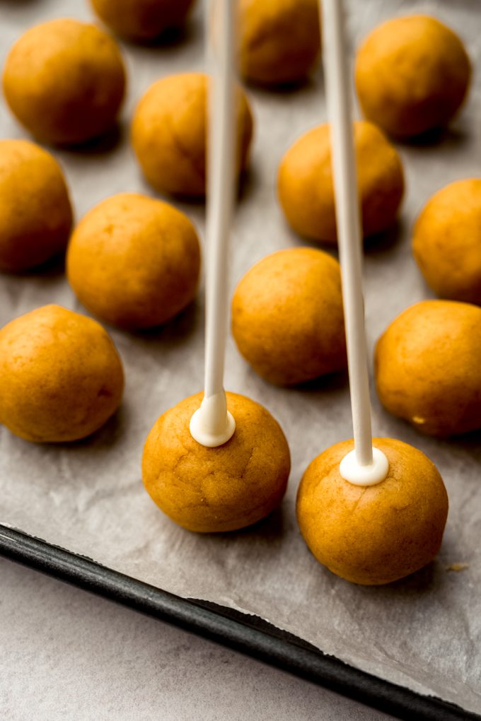 A baking sheet of cake pop balls and a couple of them have sticks in them.