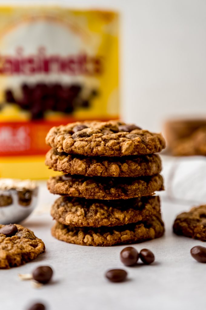 A stack of oatmeal Raisinet cookies on a surface with a bag of Raisinets in the background.