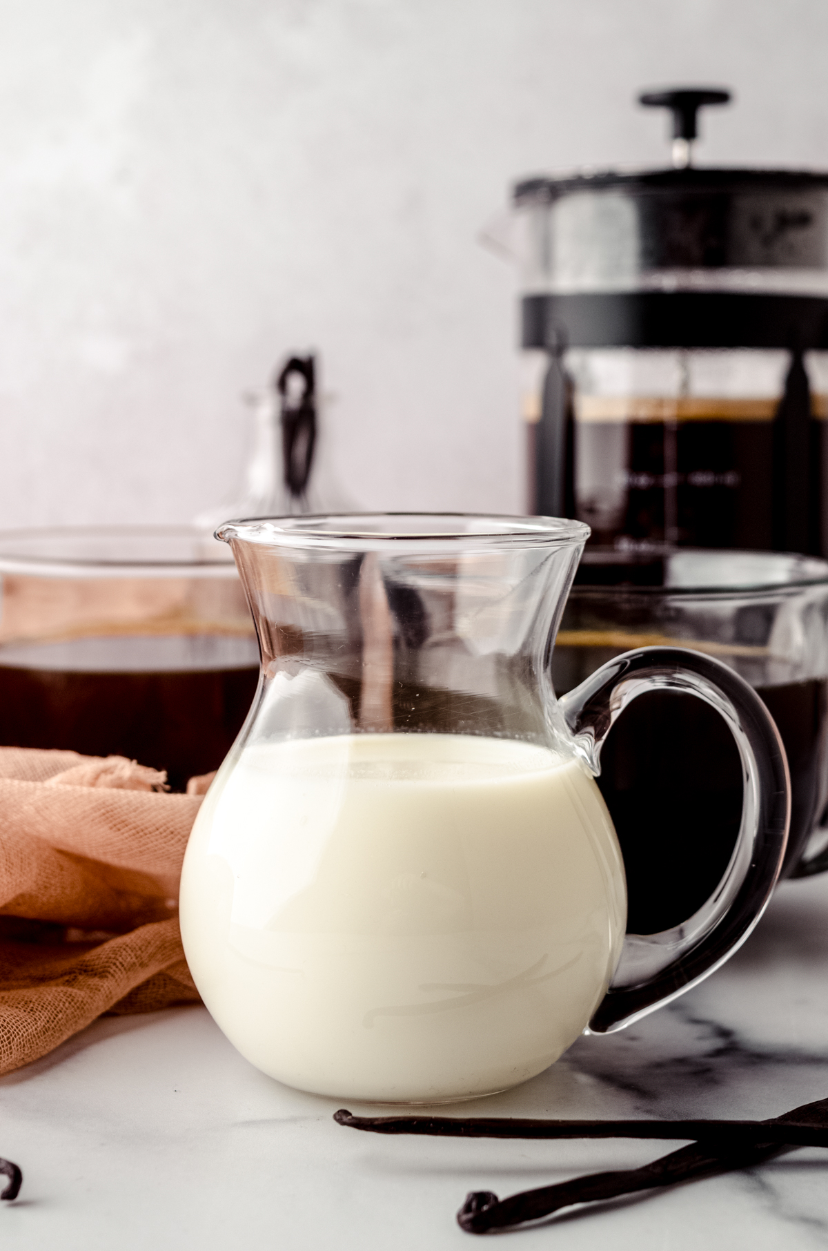 A glass pitcher with homemade French vanilla creamer in it. There are mugs of coffee and a French press in the background.