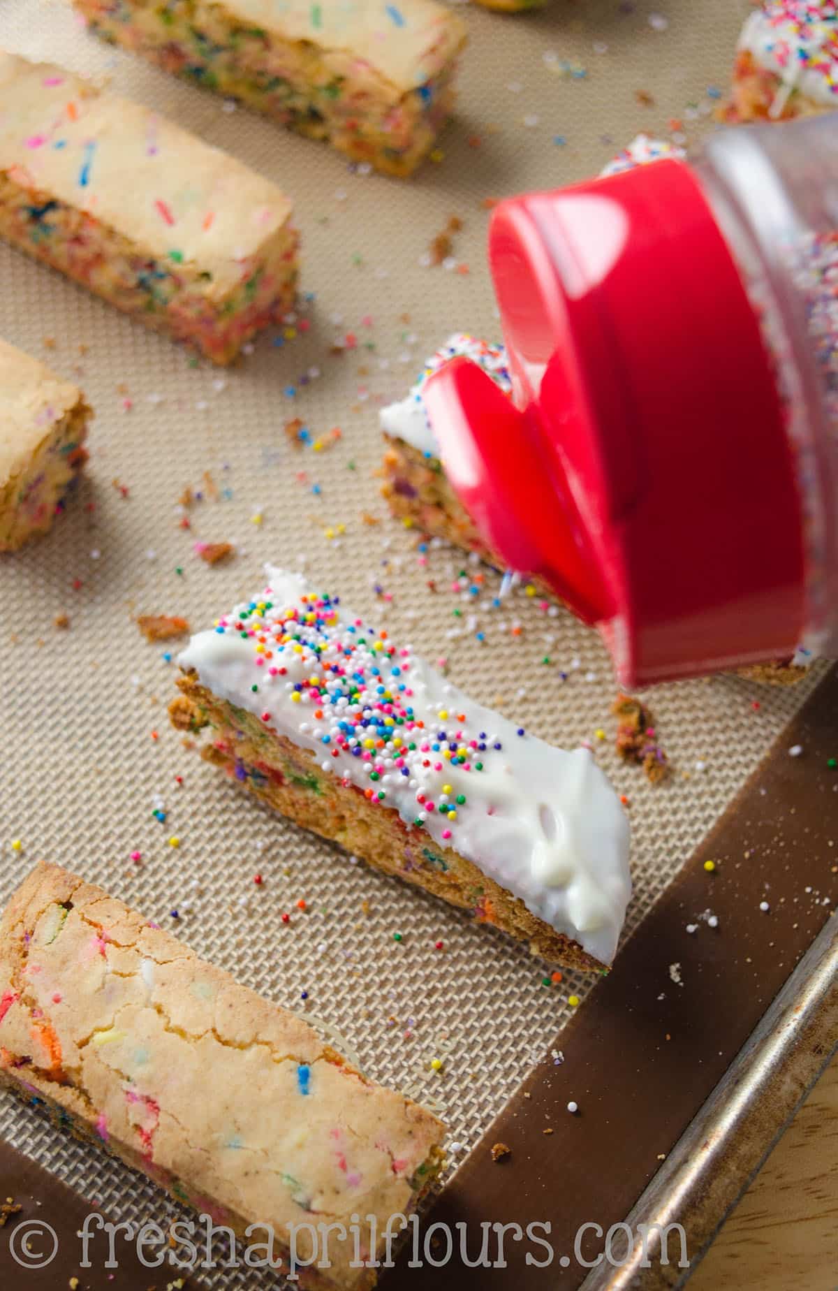 Funfetti biscotti on a baking sheet. Someone is sprinkling nonpareil sprinkles onto them.