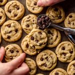 Aerial photo of a bunch of mini chocolate chip cookies with two children's hands and there is a mini cookie scoop filled with mini chocolate chips in the photo.