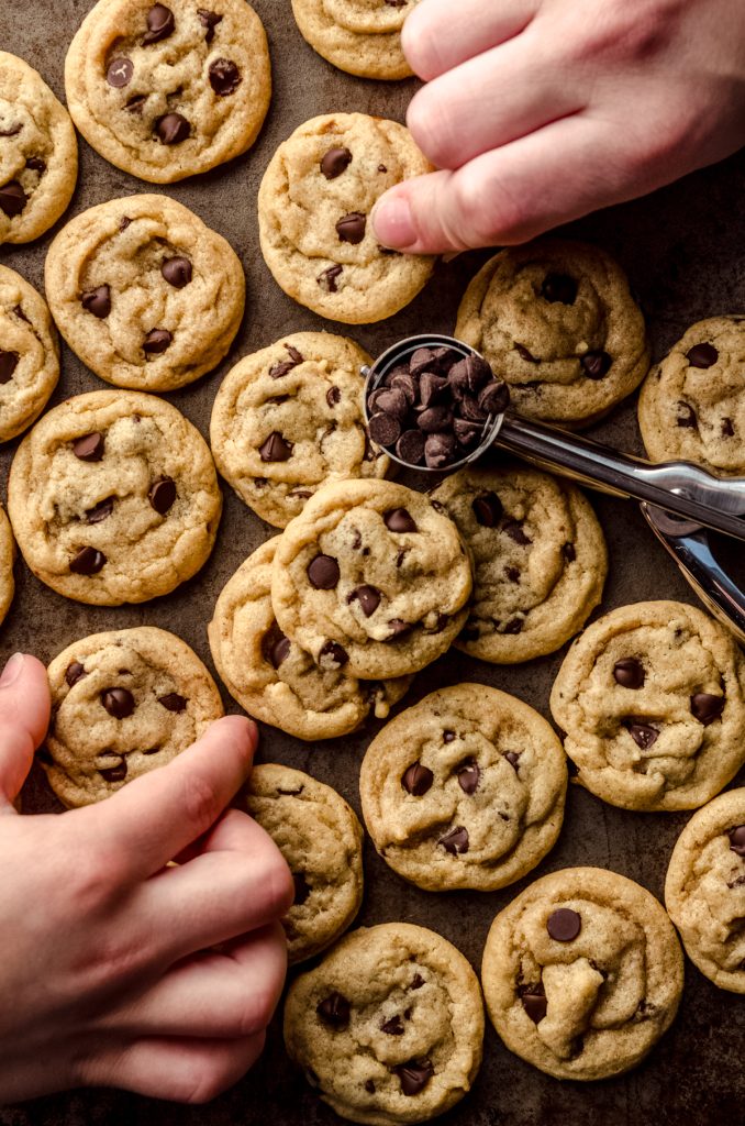 Aerial photo of a bunch of mini chocolate chip cookies with two children's hands and there is a mini cookie scoop filled with mini chocolate chips in the photo.