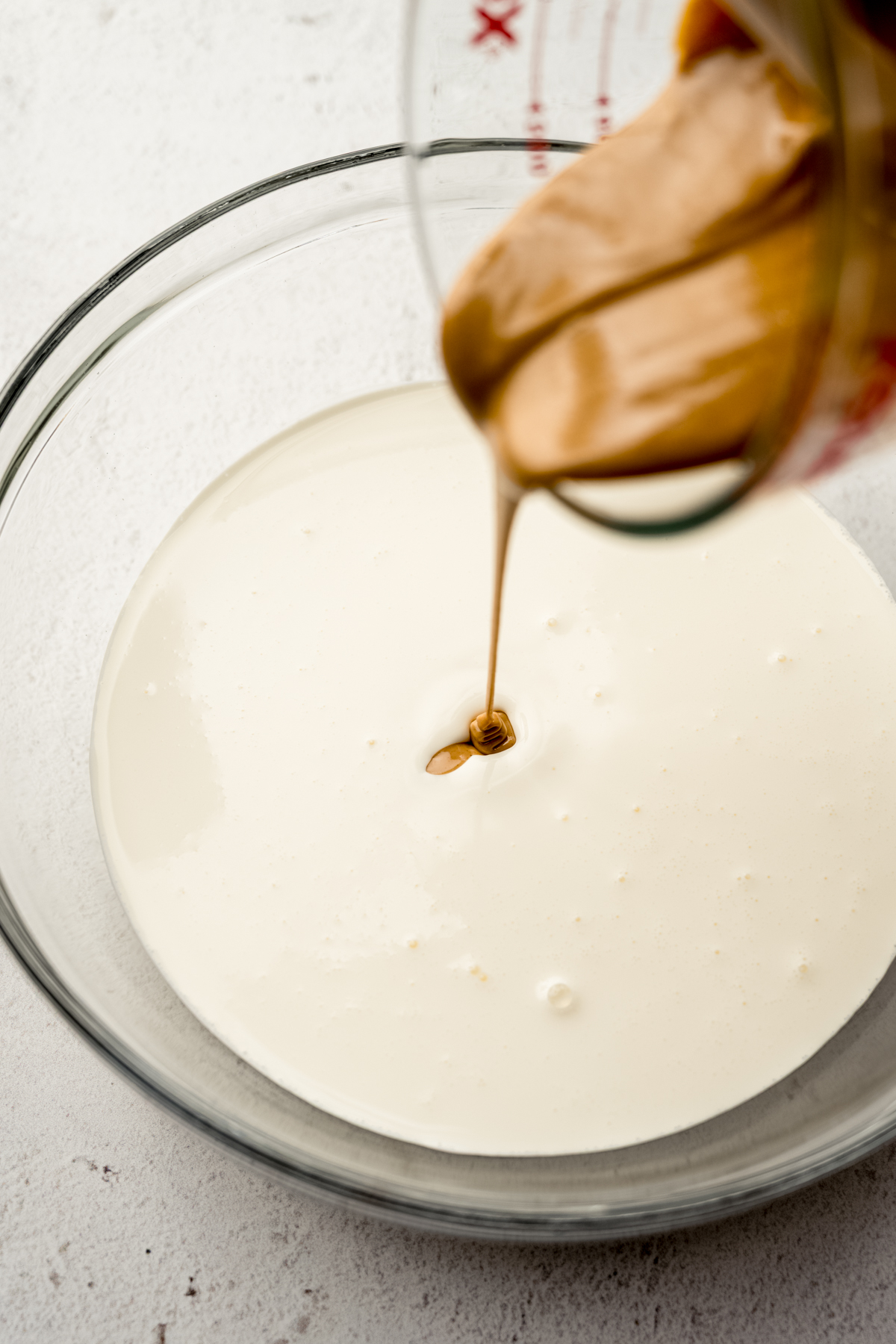 Someone is pouring melted peanut butter into a bowl of ingredients to make the base of homemade peanut butter ripple ice cream.