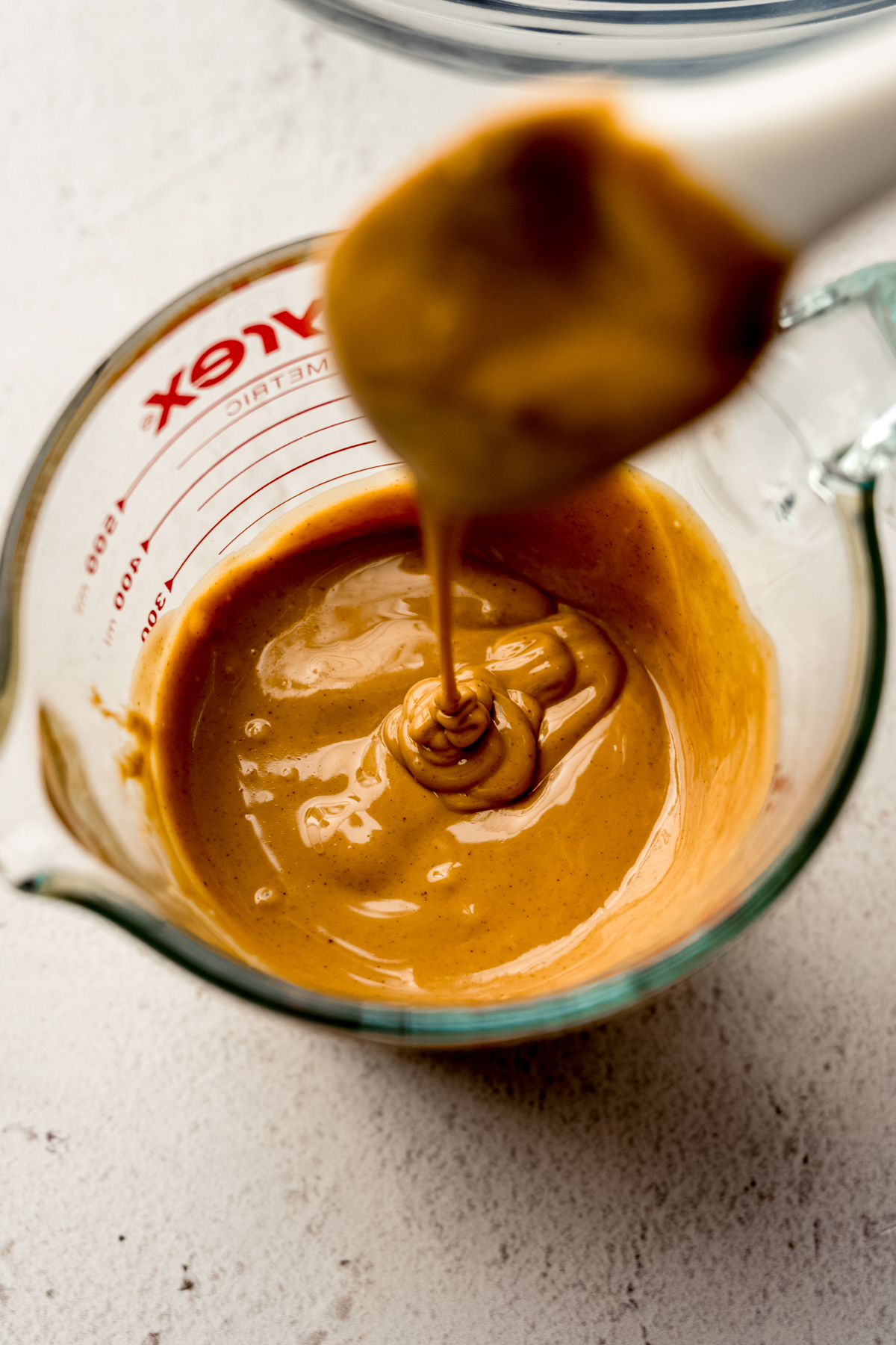 Melted peanut butter in a glass measuring cup and someone is allowing a spatula to drip into the mixture.