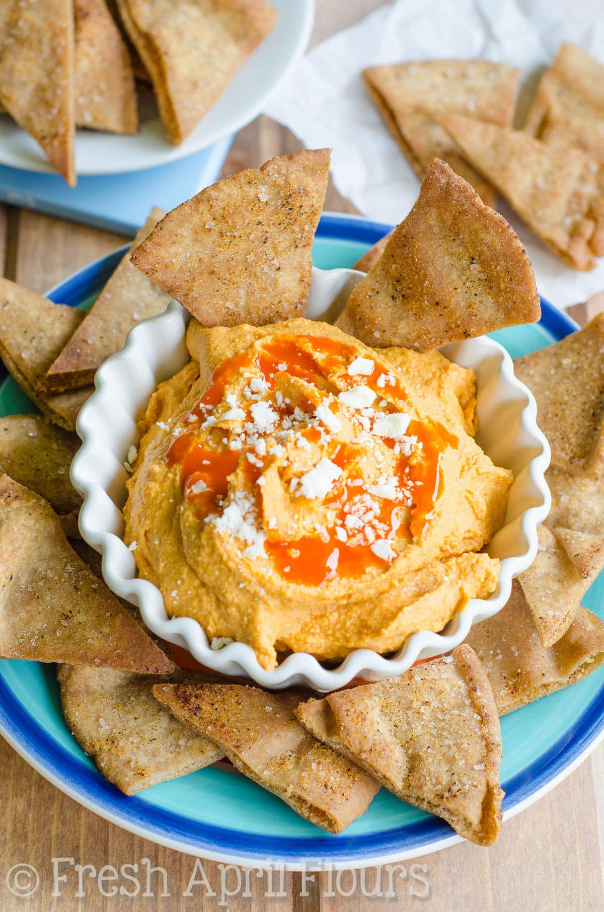 A bowl of creamy buffalo hummus on a plate with pita chips around it.