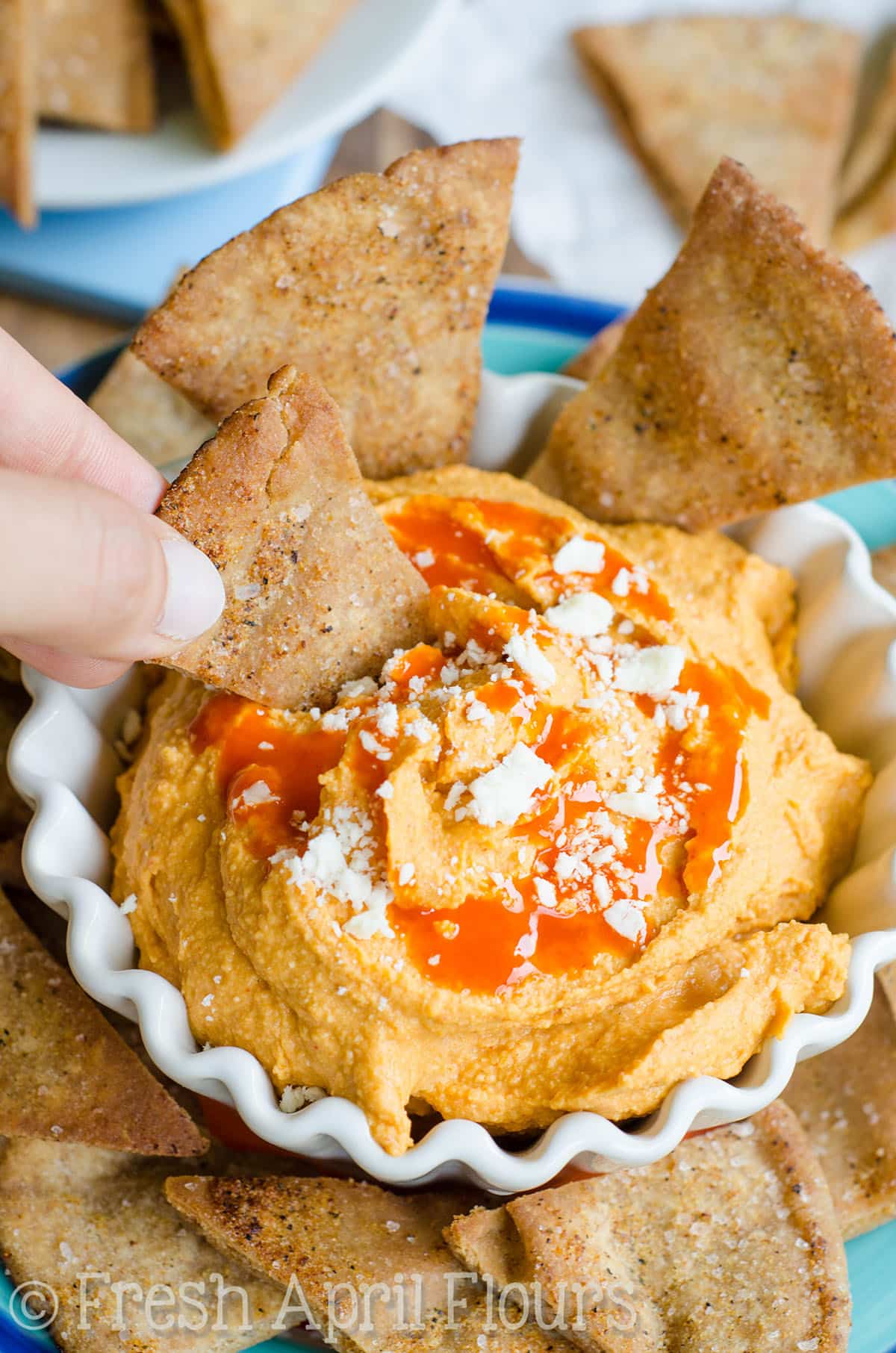 A bowl of creamy buffalo hummus on a plate with pita chips around it. A hand is dipping a pita chip into the hummus.
