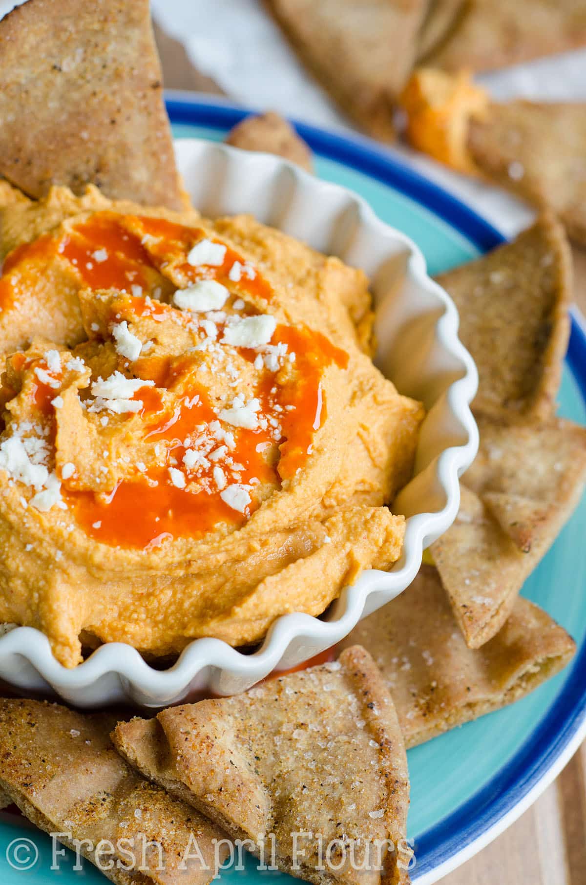 Buffalo hummus in a bowl with pita chips around it on a plate.