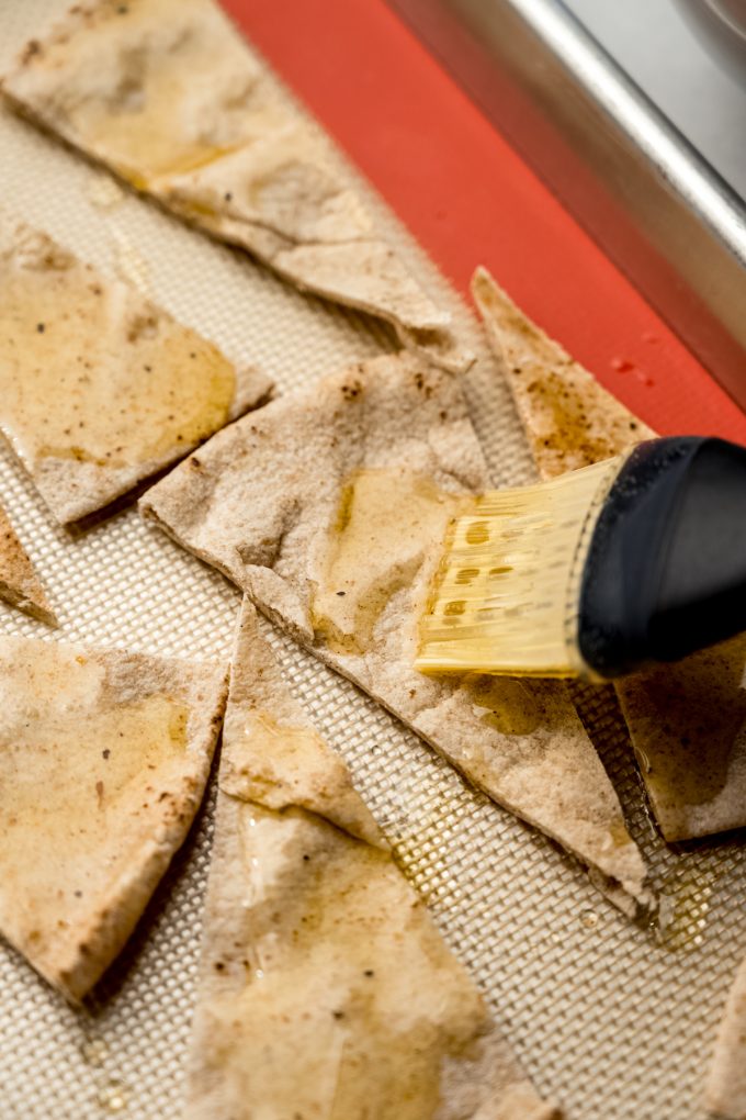 Someone is using a pastry brush to brush oil on pita bread wedges to make homemade pita chips.