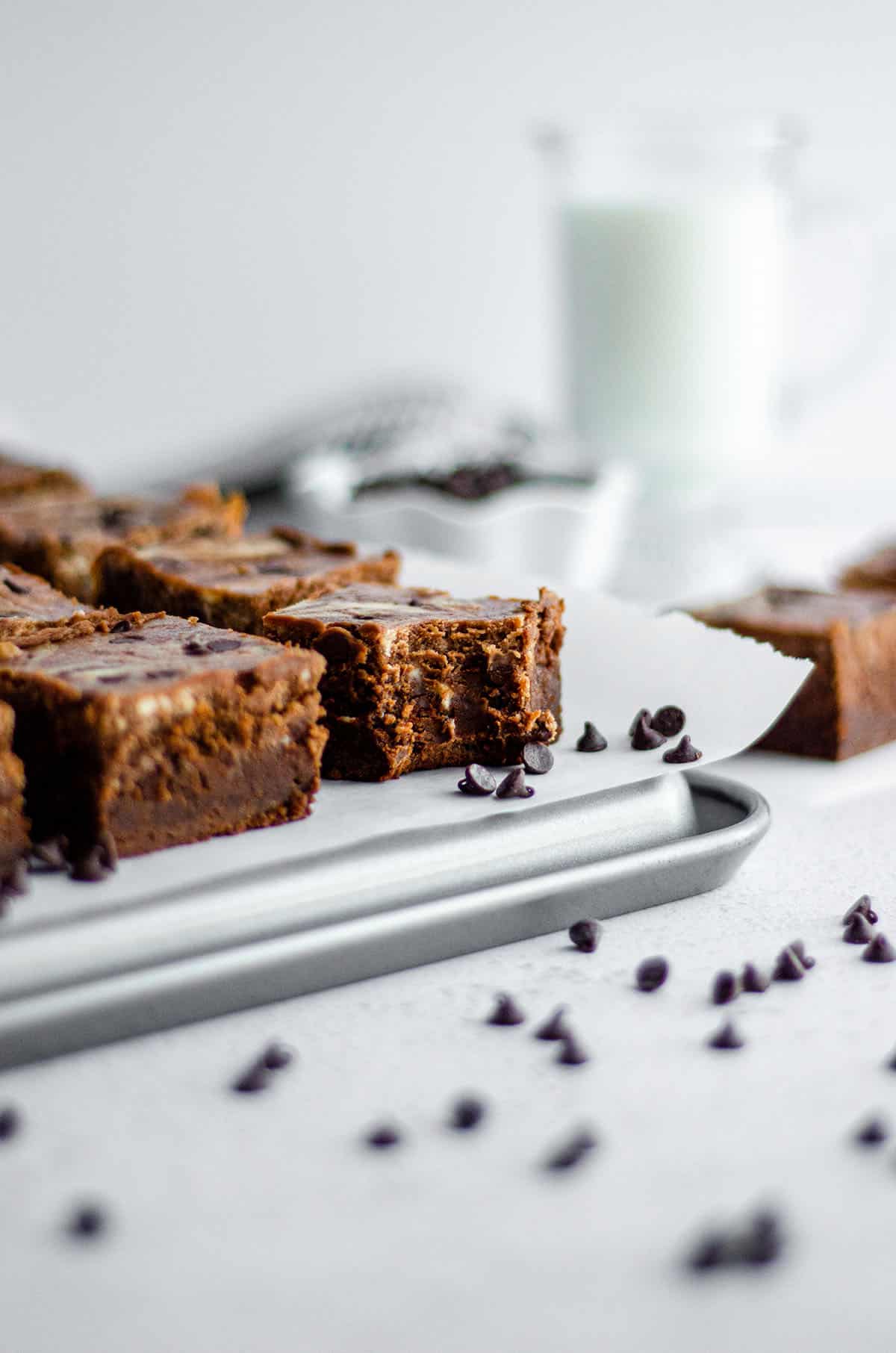 cut brownies and one with a bite taken out on parchment ready to serve with tongs, a glass of milk, and bowl of mini chocolate chips