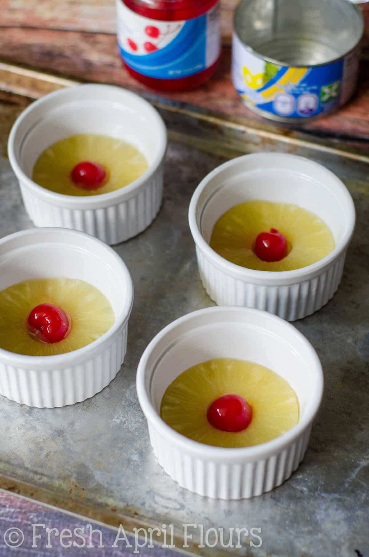 Four ramekins prepared with a pineapple ring and cherry in the middle to make mini pineapple upside down cakes.