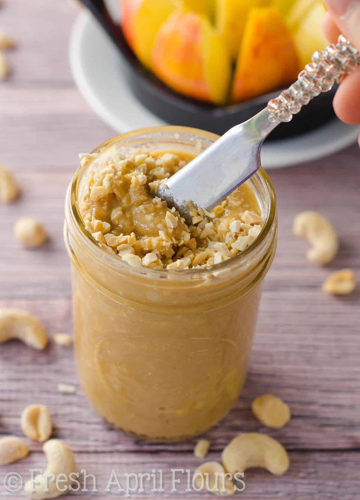 A jar of salted honey cashew peanut butter with a hand dipping a spreader into it.