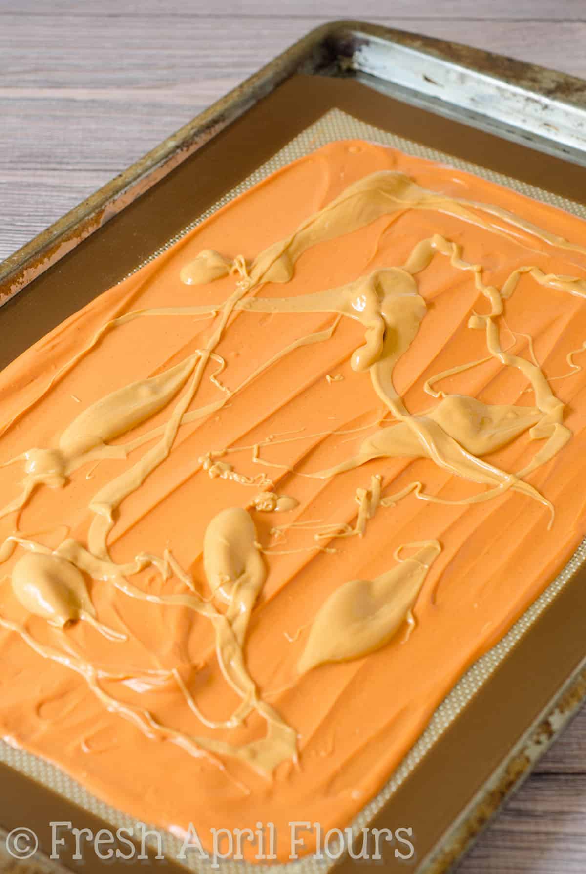 Pumpkin candy melts spread onto a baking sheet with melted butterscotch chip mixture dolloped on top.
