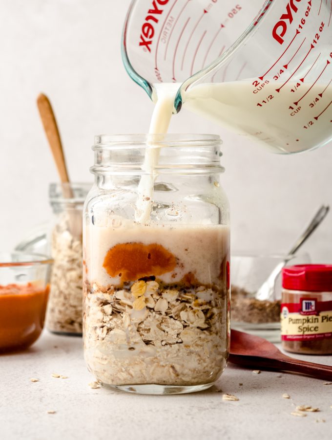 Someone is pouring milk into a Mason jar of ingredients to make pumpkin overnight oats.