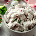 A mug full of peppermint candy cane puppy chow with festive sprinkles and Christmas decor around it.