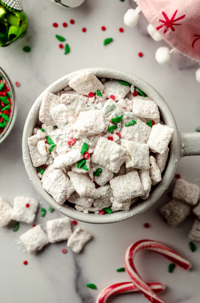 A mug full of peppermint candy cane puppy chow with festive sprinkles and Christmas decor around it.