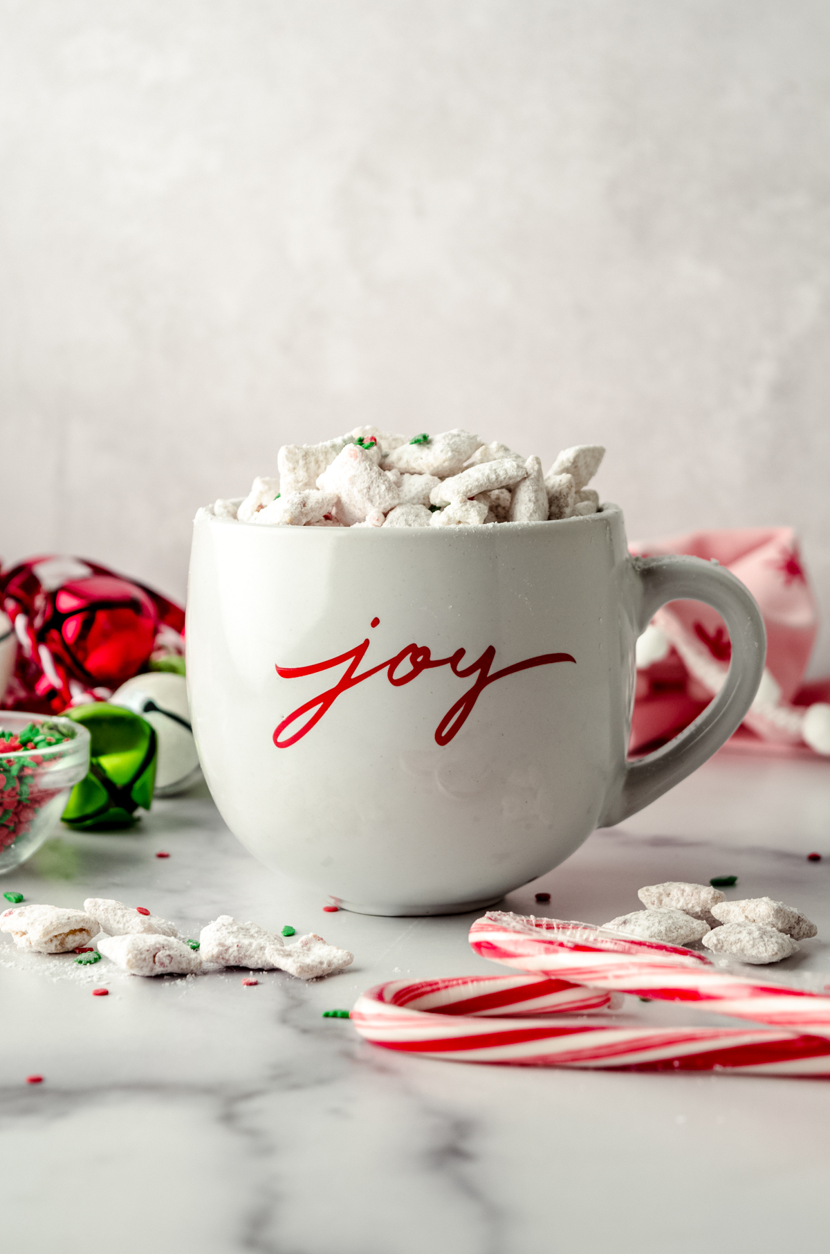 A white mug with "joy" written on it in red script with candy cane puppy chow in it with festive decor around it.