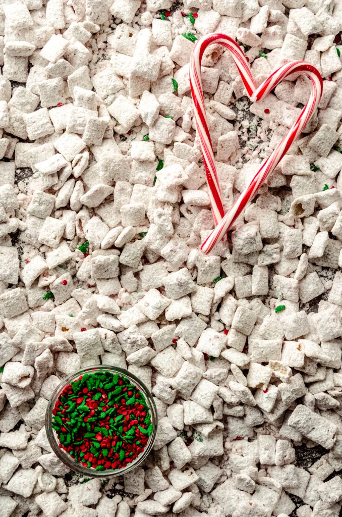 Peppermint candy cane puppy chow on a surface. There are candy canes and a bowl of festive sprinkles in the photo.