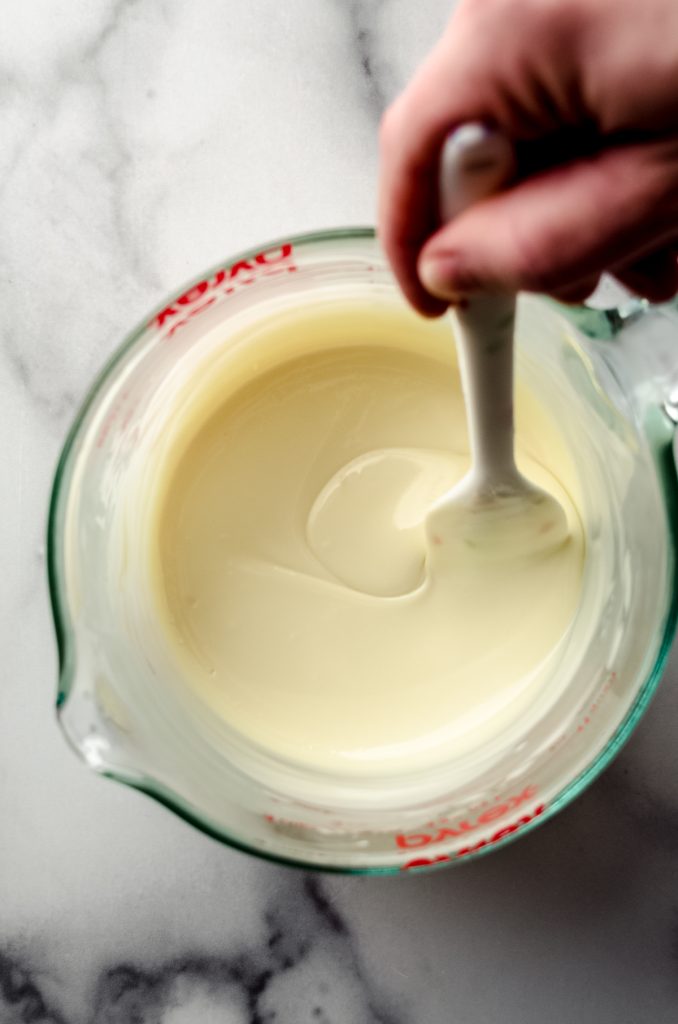 Aerial photo of someone stirring a melted white chocolate mixture in a large glass measuring cup with a spout.